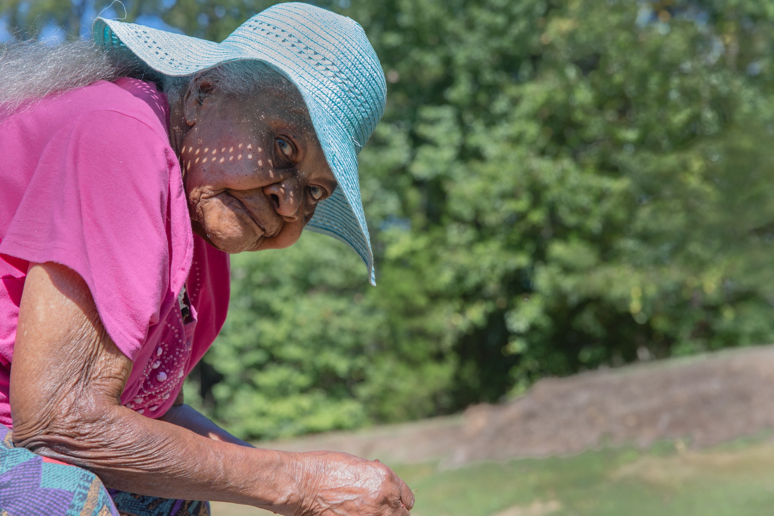 Medium close up of a 100 year old woman outside wearing a light blue sun hat and pink shirt. She is sitting perpendicular to the camer and has her head tirned to the camera lens. She has acurious and relaxed look on her face. Her eyes are light blue like her hat. This photograph is part of the To Live 10,000 Years series by Danny Goldfield.